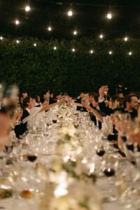 Invitados brindando en una boda de noche alrededor de una mesa larga decorada con luces cálidas y flores elegantes